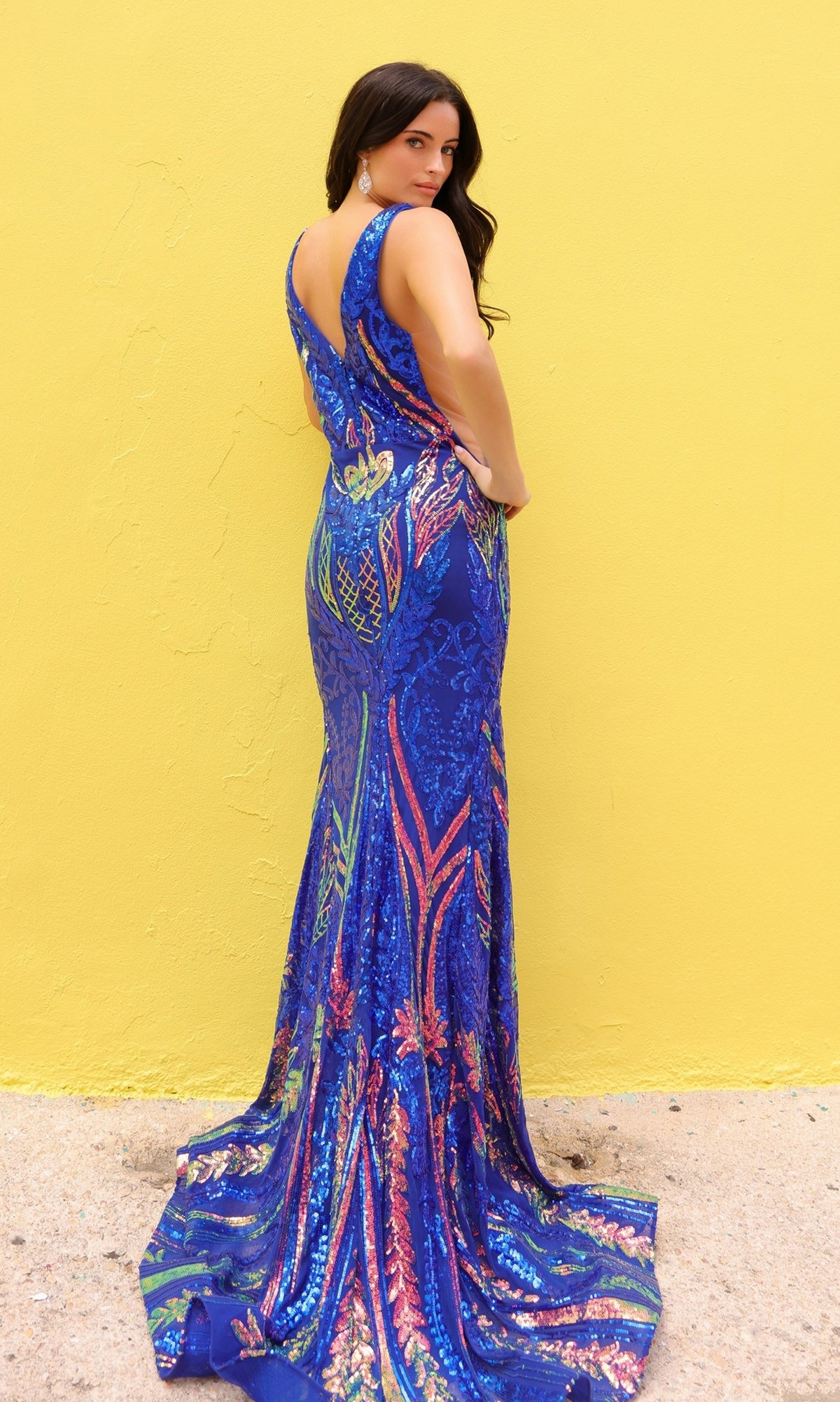 A woman in the Sheer-Sides Long Blue Sequin Prom Dress (R1402) stands against a yellow wall, gazing over her shoulder at the camera.